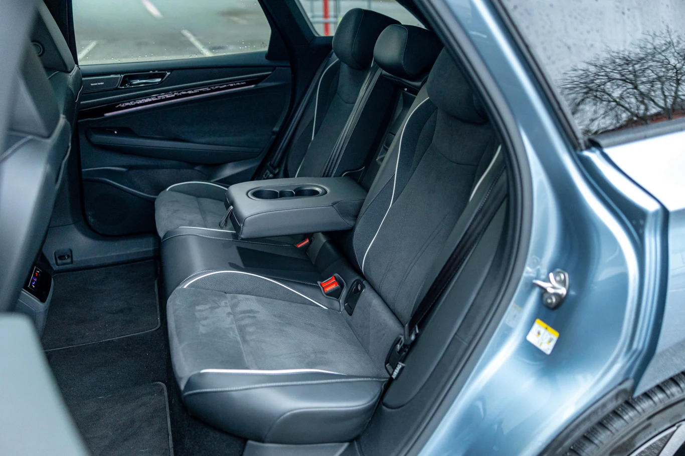 Miejsca na tylnej kanapie jest więcej niż wystarczająco Wnętrze tylnej części samochodu z dwoma wygodnymi siedzeniami wykończonymi czarną tapicerką z jasnymi przeszyciami, centralnym podłokietnikiem wyposażonym w uchwyty na kubki i widocznym fragmentem przedniego fotela.