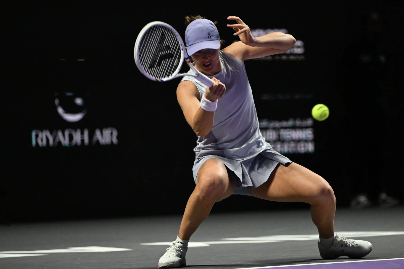 Tenisistka w jasnej sportowej sukience wykonuje efektowny manewr, próbując odbić zbliżającą się piłkę tenisową na korcie o ciemnym podłożu. W tle widoczna reklama linii lotniczych.