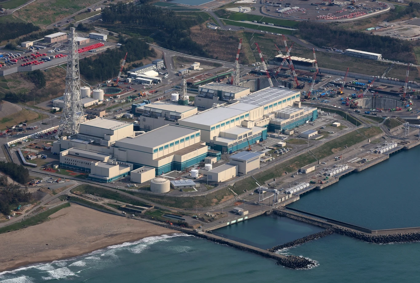 Japonia przygotowuje się do ponownego uruchomienia największej elektrowni jądrowej na świecie Kompleks budynków elektrowni jądrowej zlokalizowany nad brzegiem morza, otoczony infrastrukturą techniczną, wieżami, masztami i drogami serwisowymi, w tle widoczne tereny przemysłowe oraz miejsca parkingowe.