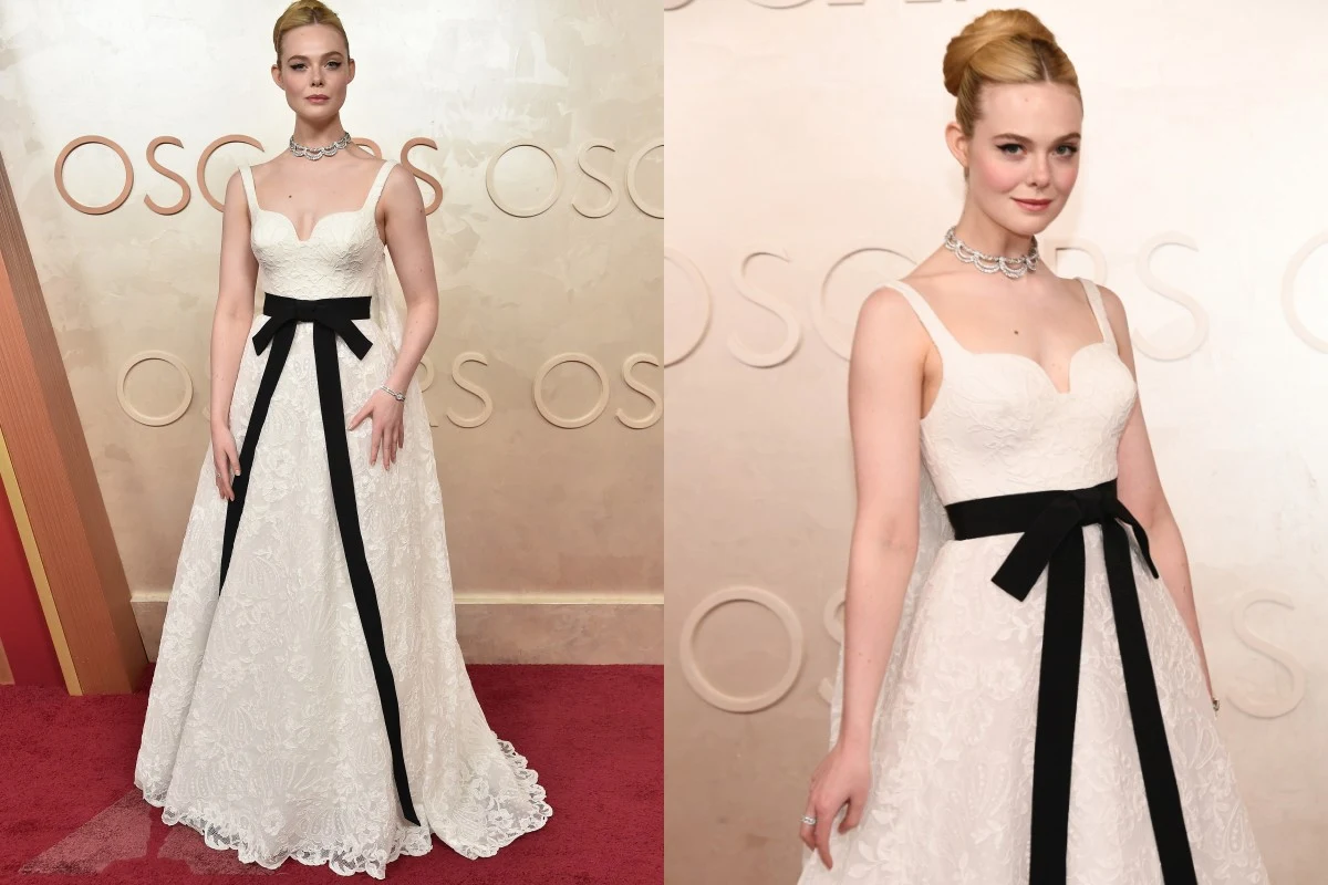 Kobieta w eleganckiej, długiej białej sukni z czarną kokardą w talii pozuje na czerwonym dywanie, na tle ściany z napisem OSCARS. Ułożone włosy, wyrazista biżuteria, stylizacja wyraźnie przygotowana na uroczyste wydarzenie.