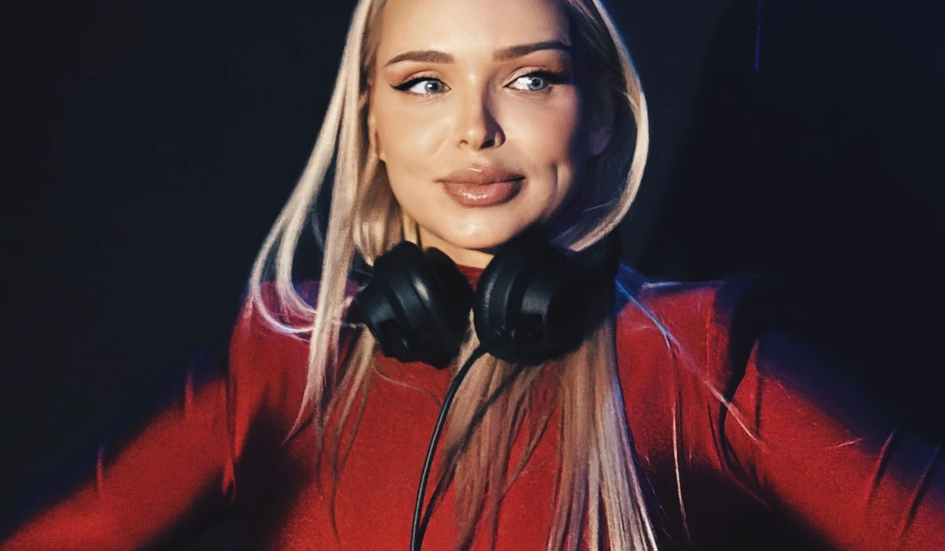 Candy Girl / Basia Hetmańska Kobieta z długimi blond włosami w czerwonym ubraniu, na szyi słuchawki DJ-skie, oświetlona z jednej strony, na ciemnym tle.