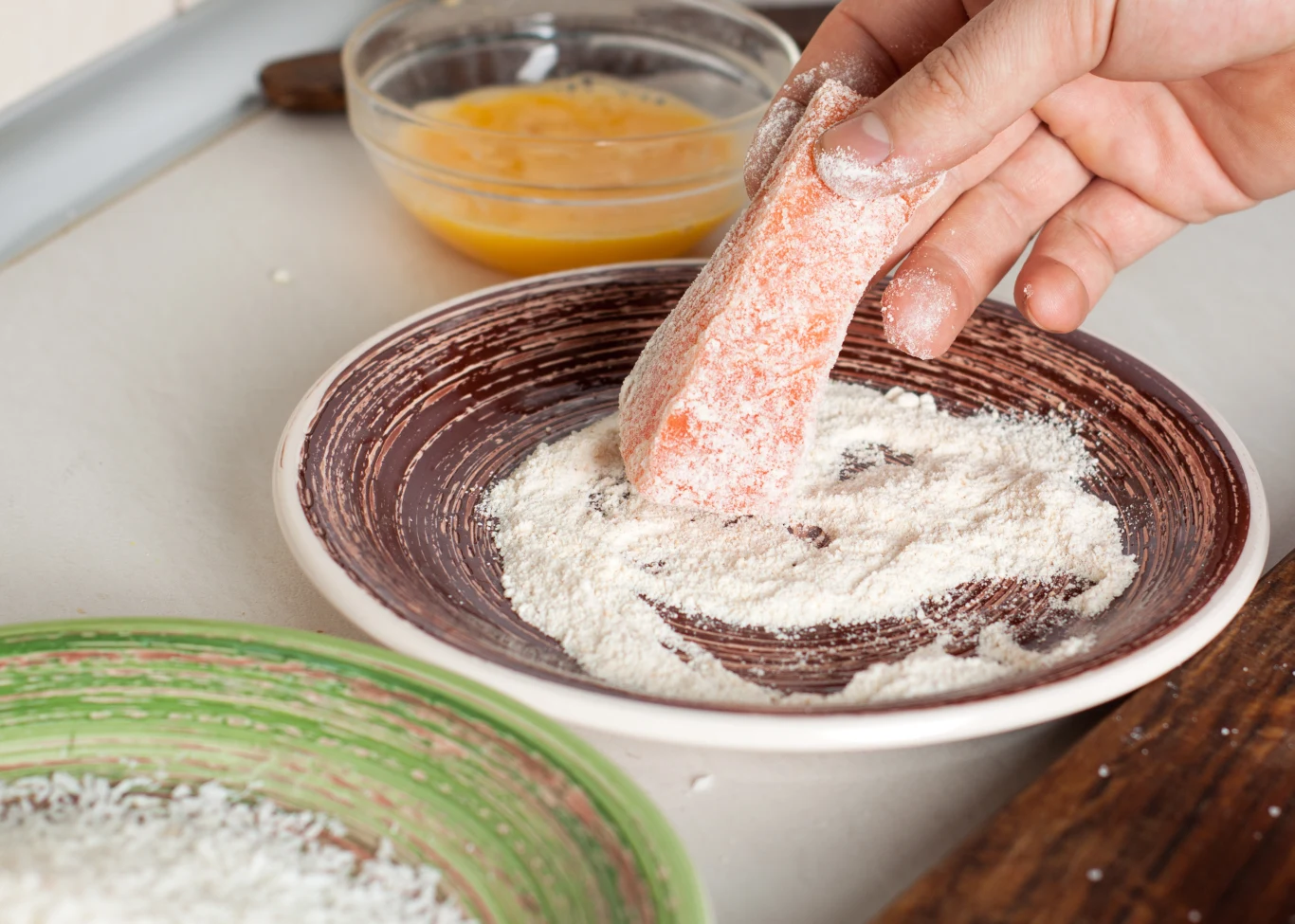 Ręka obtacza kawałek surowej ryby w mące na głębokim talerzu, w tle widoczna miska z roztrzepanym jajkiem oraz talerz z bułką tartą. Proces przygotowania do panierowania kolejnych składników.