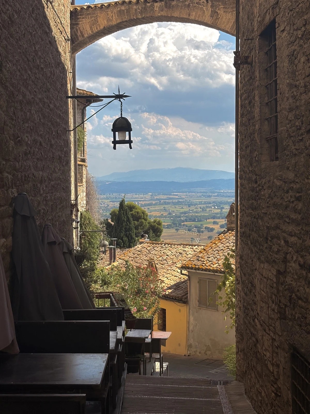 Kamienne uliczki prowadzą przez niski łuk, zza którego roztacza się widok na dachy domów, drzewa i rozległą dolinę w tle; na pierwszym planie znajdują się stoliki kawiarni ustawione wzdłuż wąskiej uliczki.