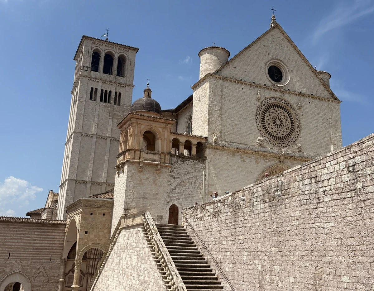 Kamienna bazylika z wysoką dzwonnicą, charakterystyczną różą na fasadzie i masywnymi schodami prowadzącymi na dziedziniec, całość otoczona jasnymi murami i utrzymana w romańskim stylu architektonicznym.