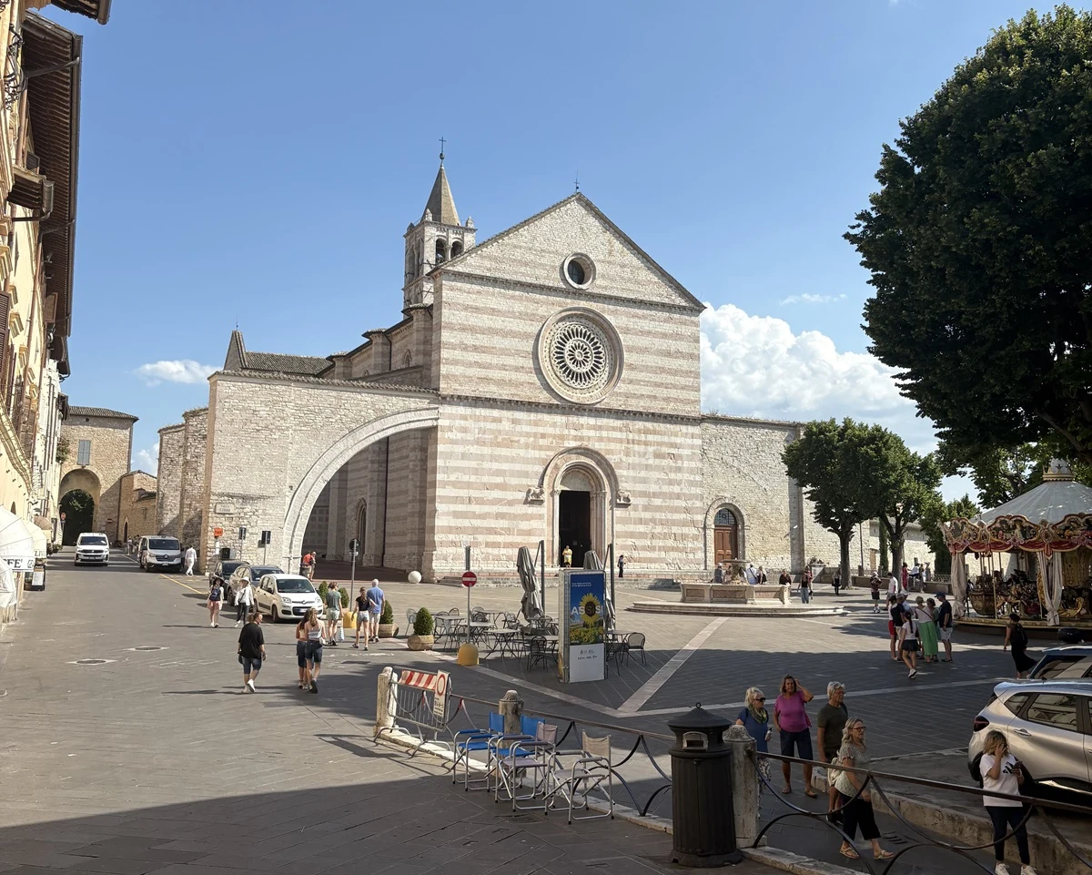 Kościół o biało-różowej fasadzie w stylu romańskim dominuje na placu miejskim. Przed świątynią widoczne są drzewa, kawiarniane stoliki, przechodnie oraz pojedyncze zaparkowane samochody. Po prawej stronie znajduje się karuzela, a wokół trwa codzienne ż...