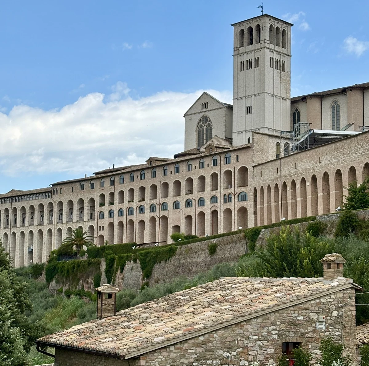 Monumentalne mury bazyliki św. Franciszka z Asyżu z charakterystyczną wieżą i arkadami rozciągają się nad zielonym wzgórzem i tradycyjnym kamiennym domem z dachówką w dolnej części zdjęcia.