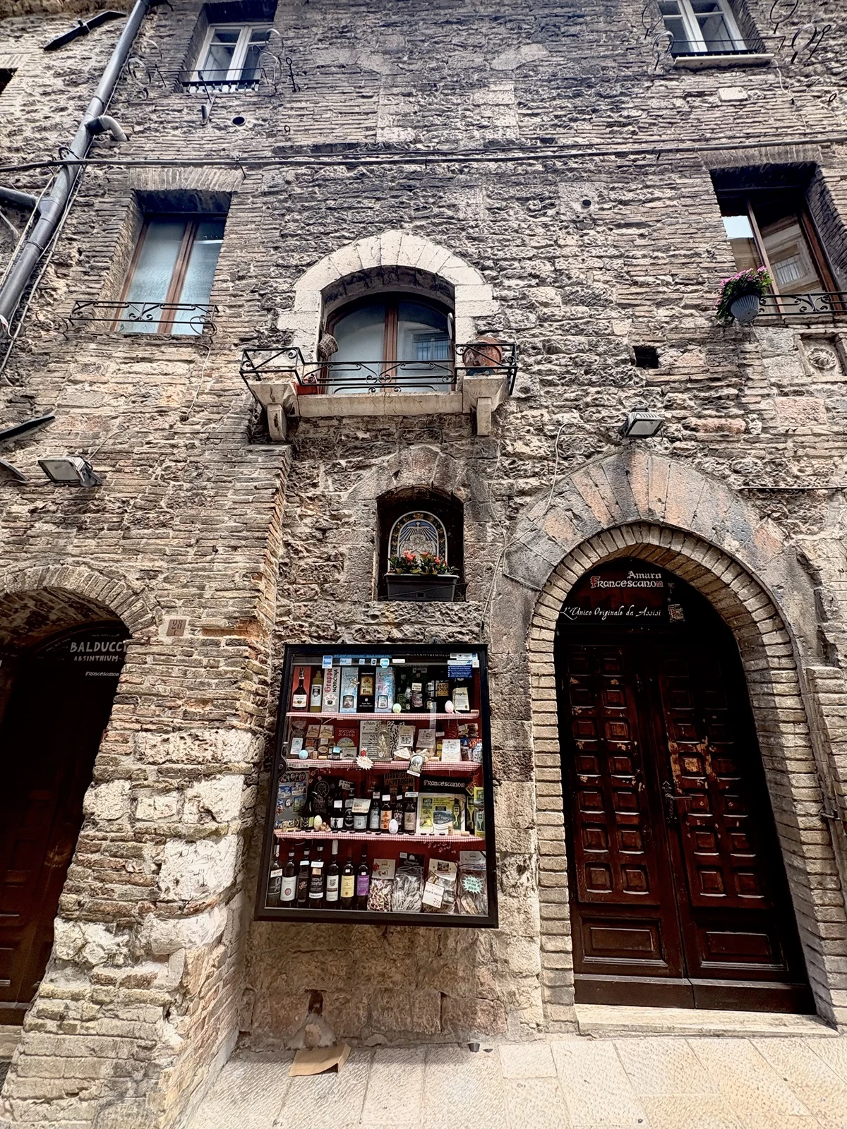 Kamienna fasada starej, wielopiętrowej kamienicy z dużymi, drewnianymi drzwiami wejściowymi oraz wystawą sklepową z alkoholami i delikatesami, okna z niewielkimi balkonami, surowa tekstura kamieni.