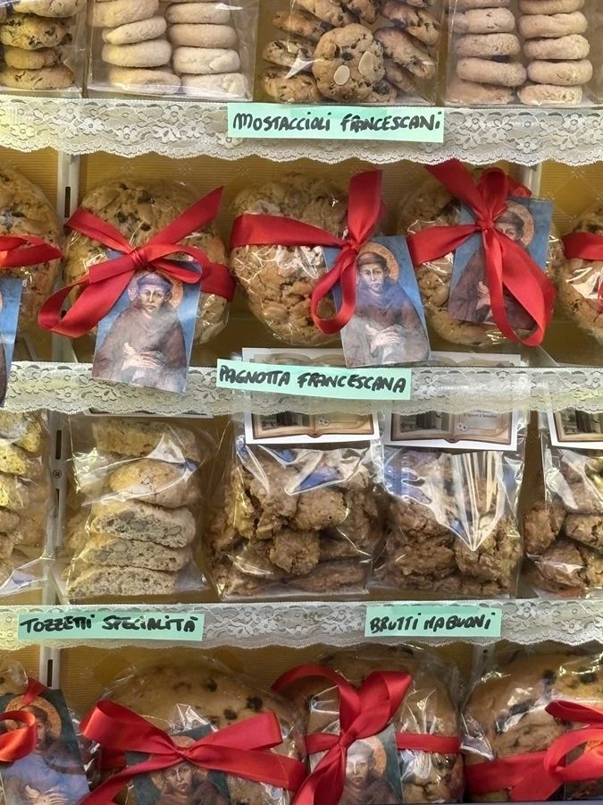 Różnorodne tradycyjne włoskie wypieki zapakowane w przezroczyste woreczki, ozdobione czerwonymi wstążkami i wizerunkami postaci religijnej, wyeksponowane na półkach z ręcznie napisanymi etykietami produktów.