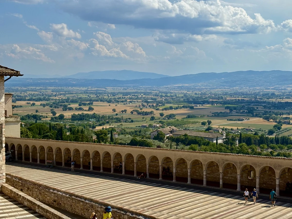 Panorama rozległej doliny z polami uprawnymi, widoczna z arkadowego tarasu z kamiennymi kolumnami; na placu spacerują pojedyncze osoby, w tle malowniczy krajobraz z pagórkami oraz rozproszonymi zabudowaniami.
