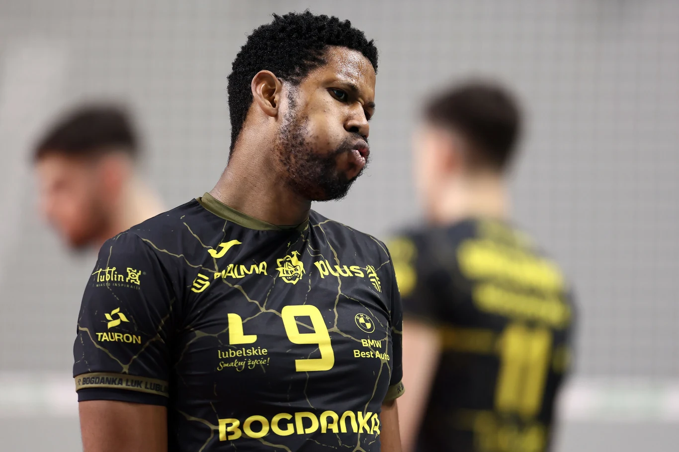 Siatkarz w czarno-żółtym stroju sportowym widoczny na pierwszym planie podczas meczu, z wyraźnym wyrazem skupienia lub rozczarowania na twarzy, w tle rozmazane postaci innych zawodników.