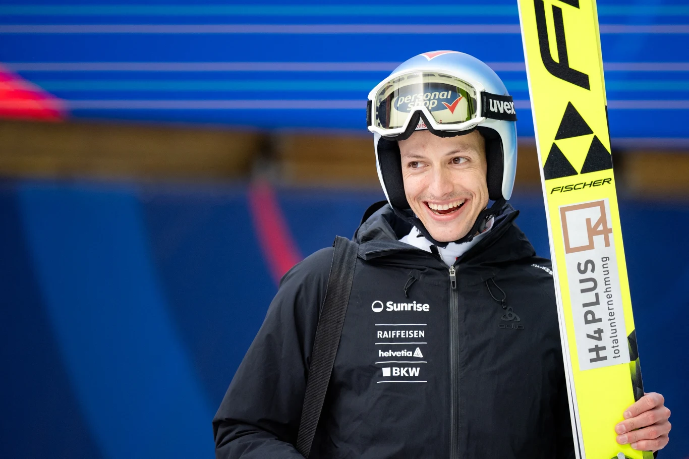 Uśmiechnięty skoczek narciarski ubrany w sportowy kombinezon i kask ochronny trzyma w dłoni narty oznaczone logo producenta, na tle niebieskiego obiektu sportowego.