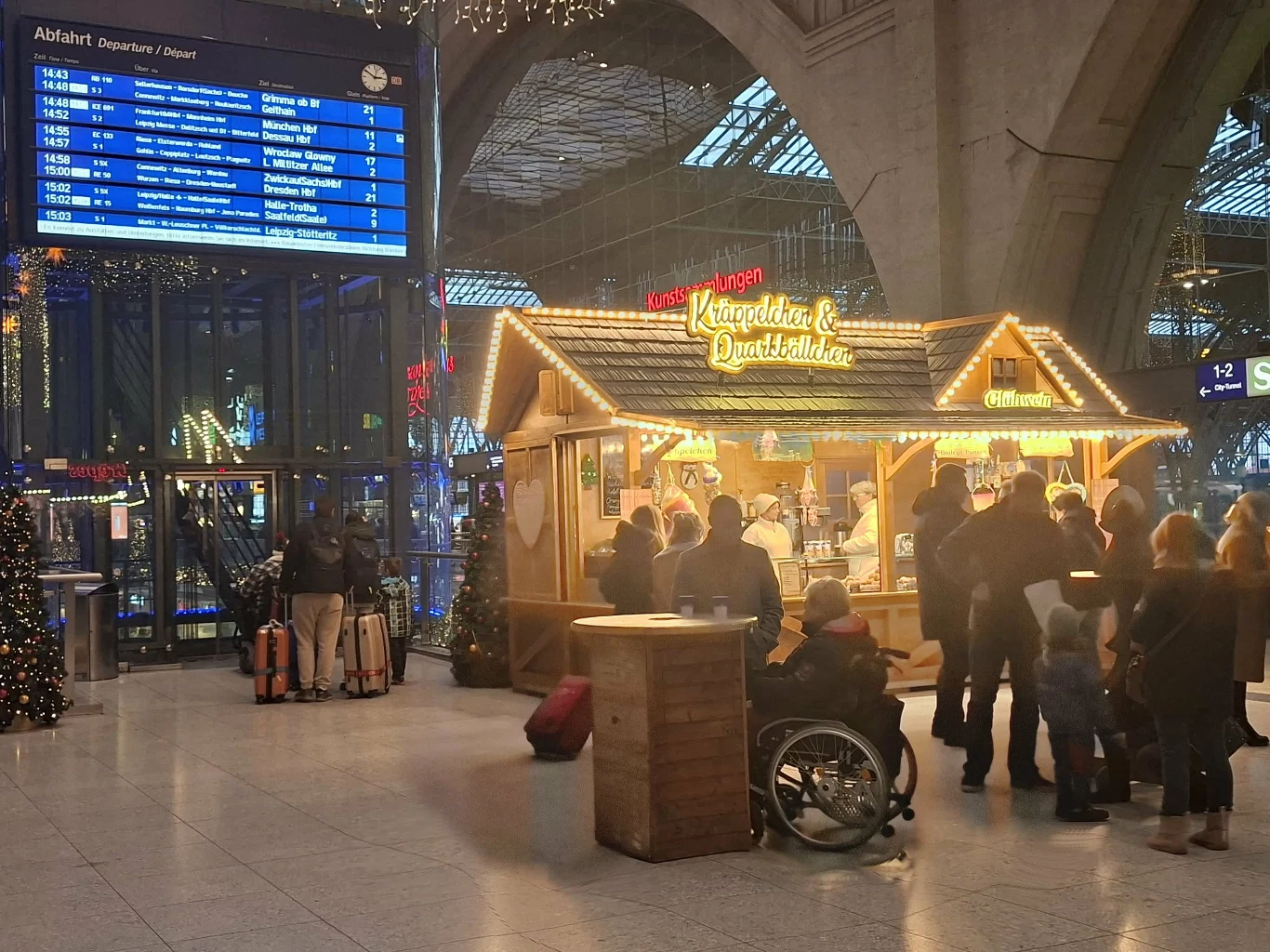 Jarmarkowa budka ozdobiona świątecznymi światełkami otoczona grupą osób w dużej hali dworca, w tle cyfrowy rozkład jazdy pociągów oraz choinki bożonarodzeniowe, jedna osoba na wózku inwalidzkim.