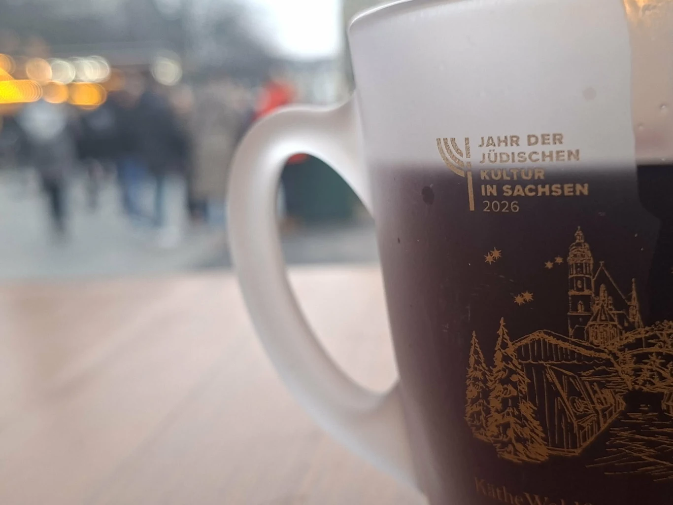 Szklanka z ciemnym napojem ustawiona na drewnianym stole, na szklance znajduje się złoty nadruk z napisem informującym o Roku Kultury Żydowskiej w Saksonii w 2026 roku, w tle rozmyte sylwetki ludzi na miejskim deptaku i nieostre światła świątecznego ki...