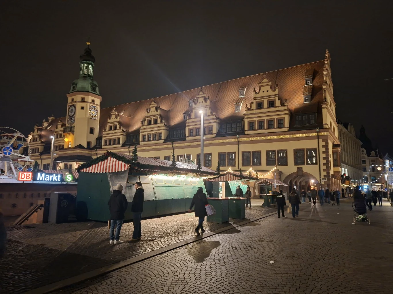 Zabytkowy budynek ratusza z charakterystycznym zegarem oświetlony nocą, na pierwszym planie jarmarczne stragany oraz spacerujący ludzie.