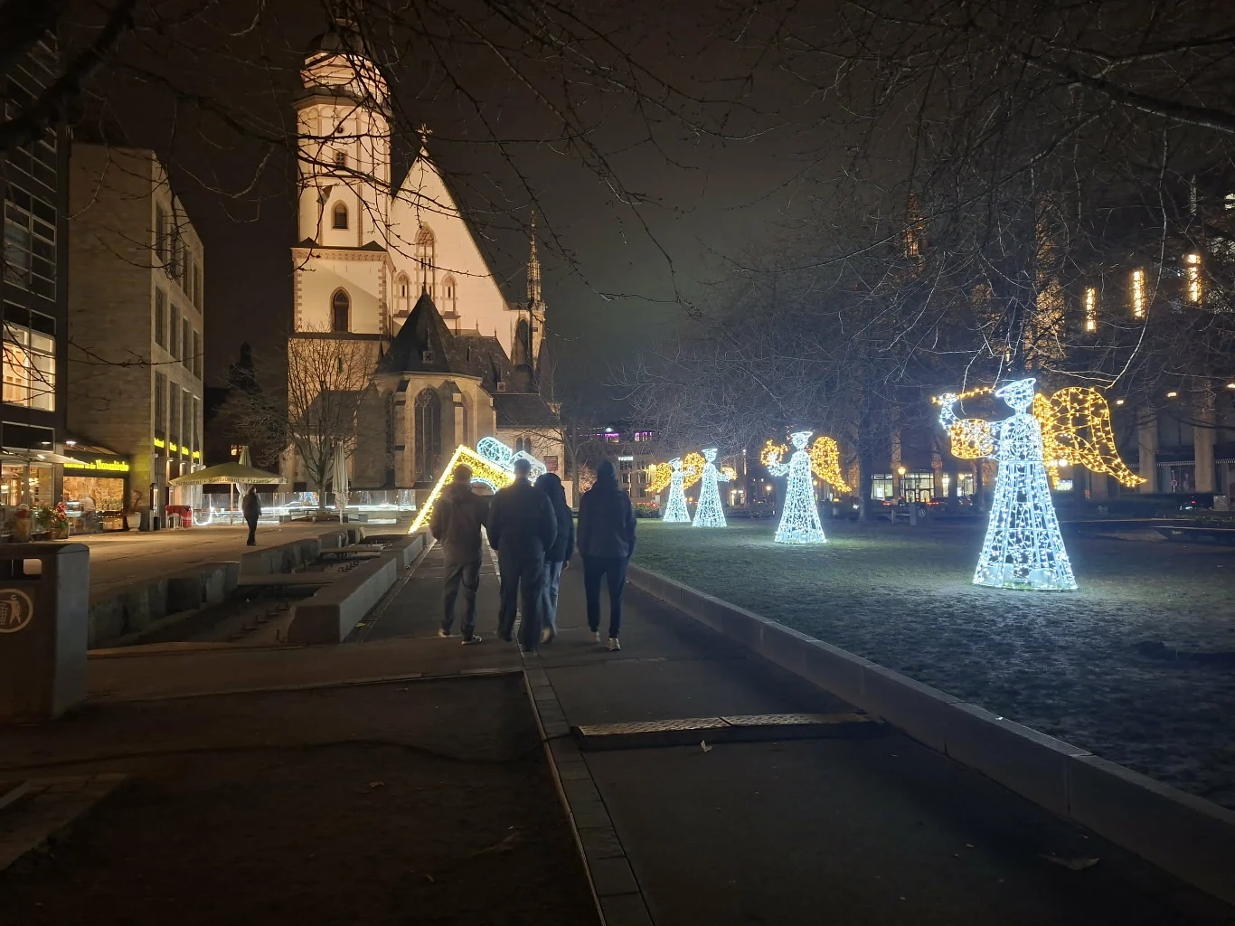 Oświetlone instalacje świetlne w kształcie aniołów ustawione wzdłuż alei nocą, w tle widoczny kościół oraz grupa osób spacerujących obok dekoracji na tle miejskiego krajobrazu.