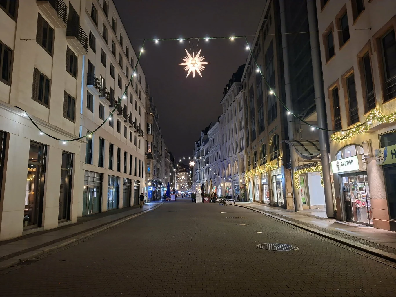 Opustoszała ulica miasta oświetlona dekoracjami świątecznymi, w tym zawieszoną gwiazdą nad jezdnią oraz girlandami światełek na elewacjach budynków, sklepy i kamienice po obu stronach ulicy.