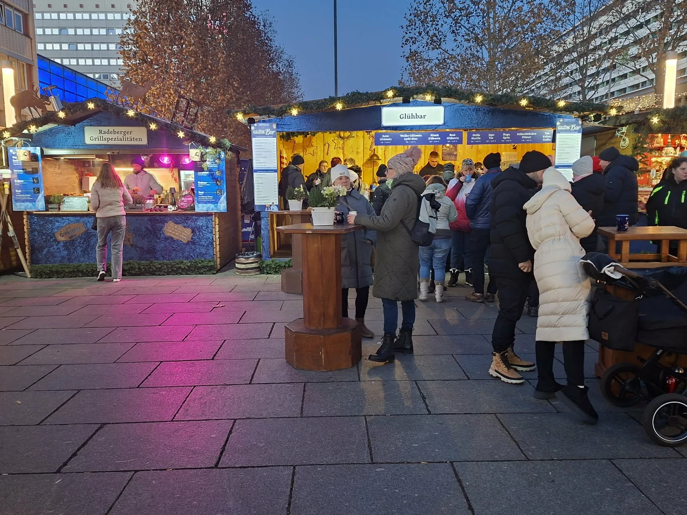 Ludzie stojący w kolejce do świątecznych stoisk gastronomicznych na tle wieczornego, miejskiego krajobrazu. Ubrani w ciepłe kurtki mieszkańcy korzystają z wysokich stojących stolików. W tle jasne dekoracje i światła jarmarku.