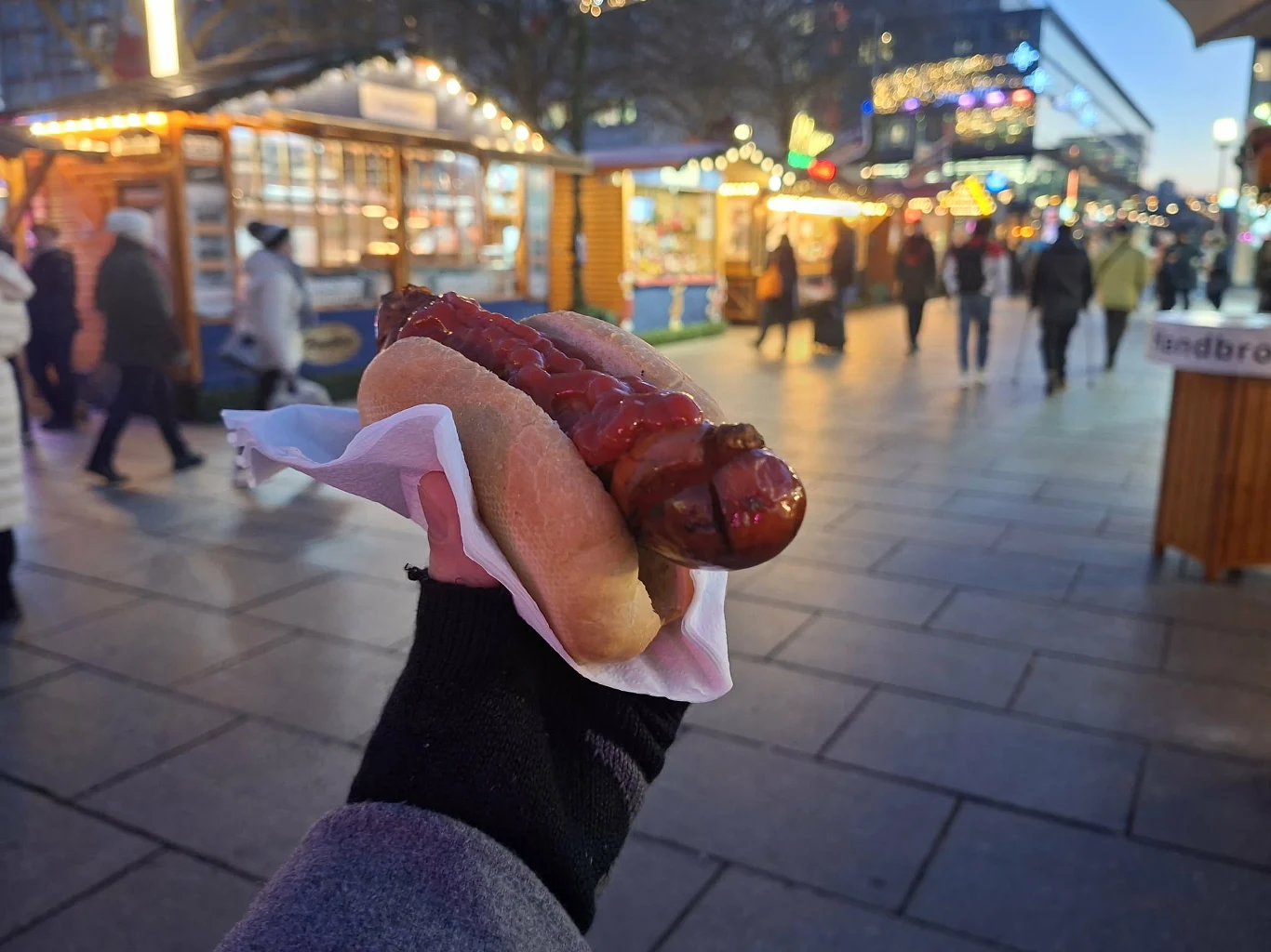 Ręka trzymająca hot doga z grillowaną kiełbasą i sosem, na tle targu świątecznego z oświetlonymi budkami i spacerującymi ludźmi.