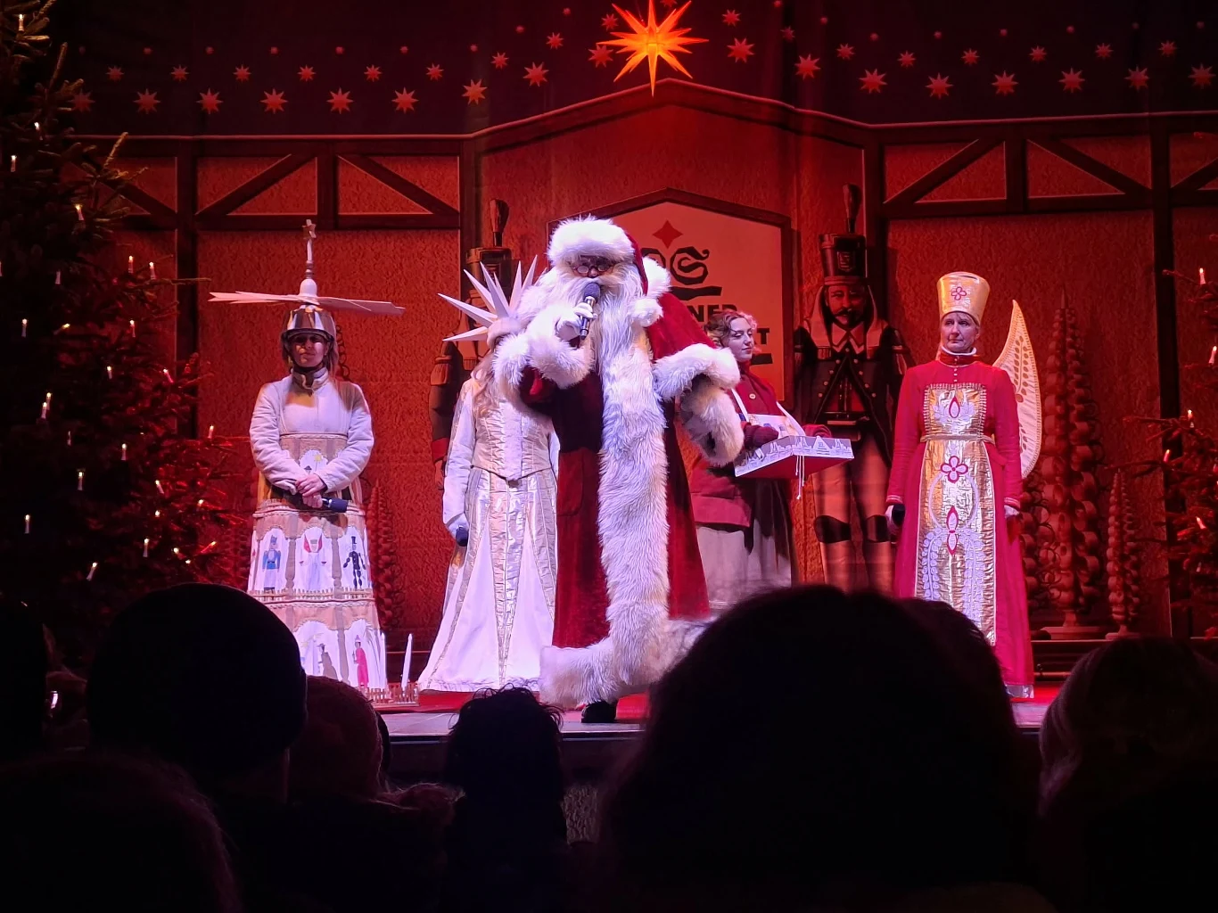 Postacie w strojach świątecznych i bajkowych stoją na oświetlonej scenie w otoczeniu dekoracji bożonarodzeniowych; centralnie Mikołaj obejmuje postać kobiety ubraną w białą suknię i koronę, obok stoją dwie inne osoby w kostiumach przypominających trady...