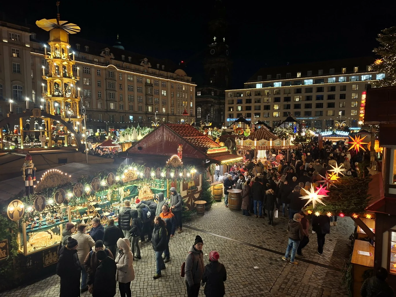 Tłum ludzi przechadza się po rozświetlonym targu świątecznym otoczonym jasnymi, kolorowymi straganami i dekoracjami, w tle zabytkowe budynki i wielka podświetlana piramida bożonarodzeniowa.