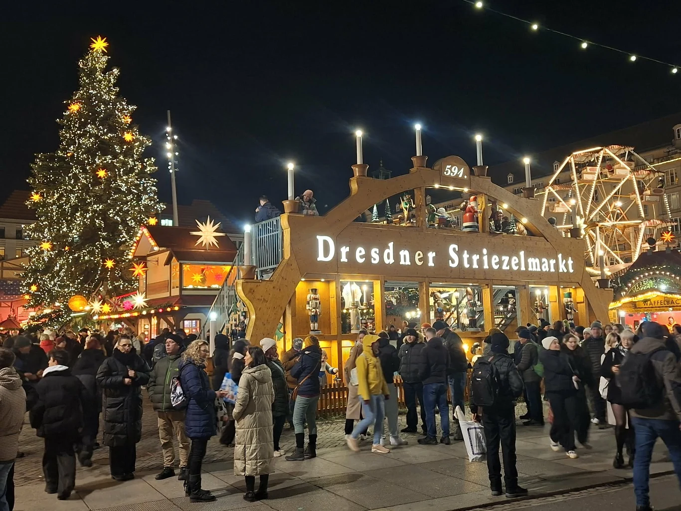 Tłum ludzi spacerujący po jarmarku bożonarodzeniowym w Dreźnie, ozdobiona choinka, iluminacje świąteczne, jasne światła i stoiska tworzą świąteczny klimat.
