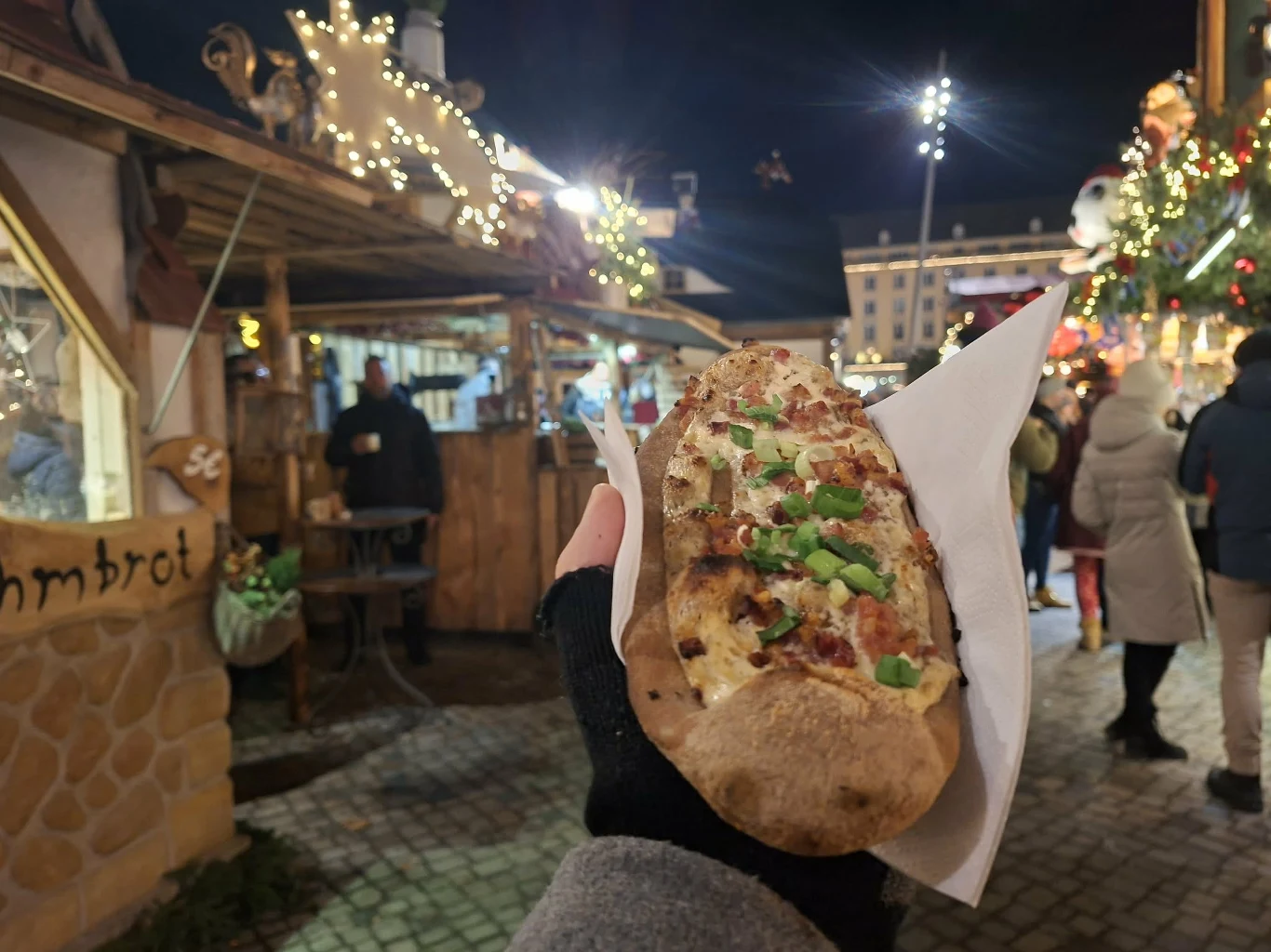 Chrupiąca bułka z nadzieniem, posypana świeżymi warzywami trzymana w ręce, w tle nastrojowy kiermasz świąteczny z oświetlonymi stoiskami i spacerującymi ludźmi.