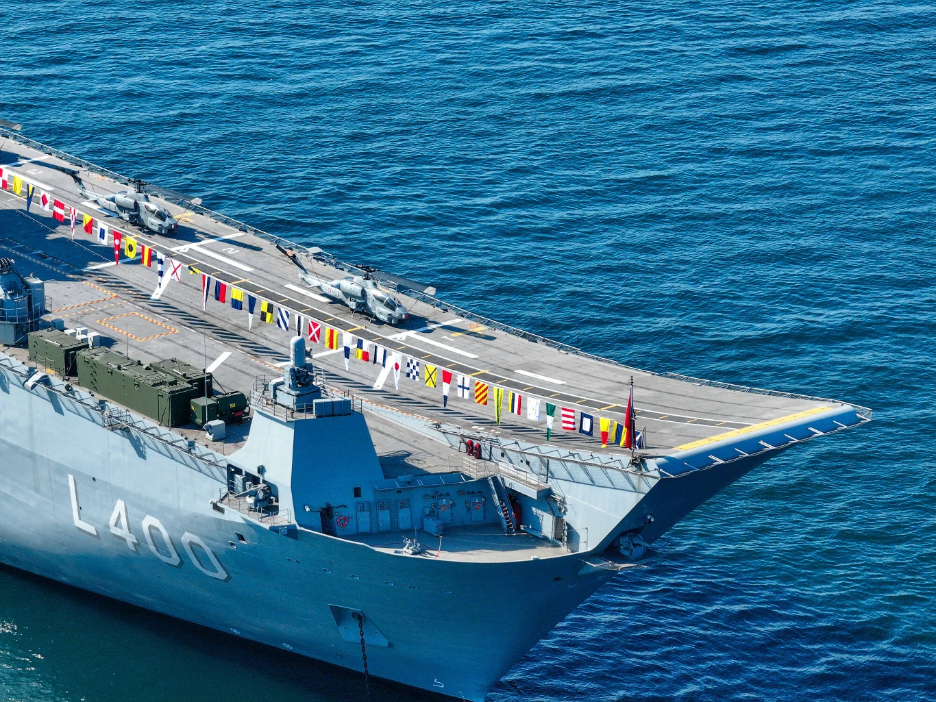Nowoczesny okręt wojskowy z numerem L400, na którego pokładzie znajdują się dwa helikoptery oraz liczne flagi sygnałowe rozmieszczone wzdłuż krawędzi. Statek znajduje się na spokojnej wodzie, a jego pokład jest uporządkowany i wyraźnie przygotowany do ...