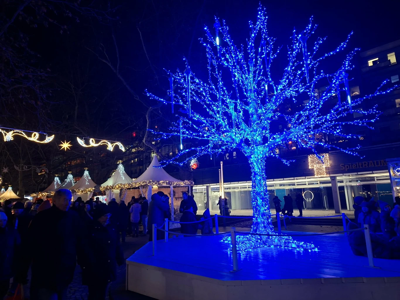 Jasno oświetlone niebieskie drzewo pokryte świątecznymi lampkami na tle jarmarku, wokół którego zgromadzili się ludzie podziwiający dekoracje, w tle widoczne białe namioty oraz świąteczne iluminacje.