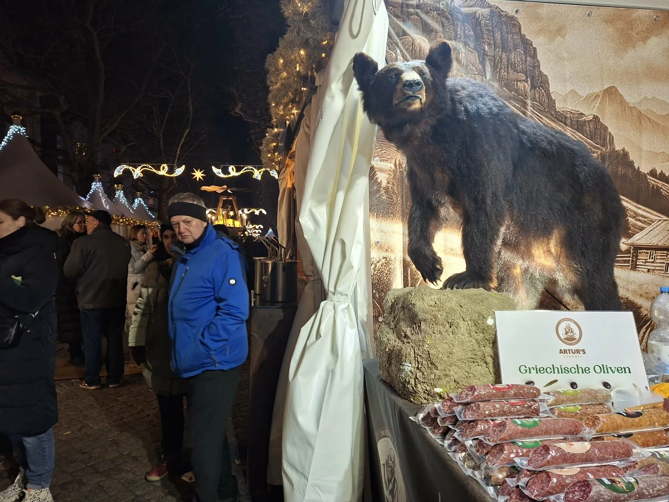 Stoisko na jarmarku nocą z wypchanym niedźwiedziem ustawionym na podwyższeniu, a przed nim ekspozycja wędlin i produktów spożywczych. Po lewej stronie widoczna grupa osób spacerujących pomiędzy stoiskami i świąteczne dekoracje świetlne w tle.