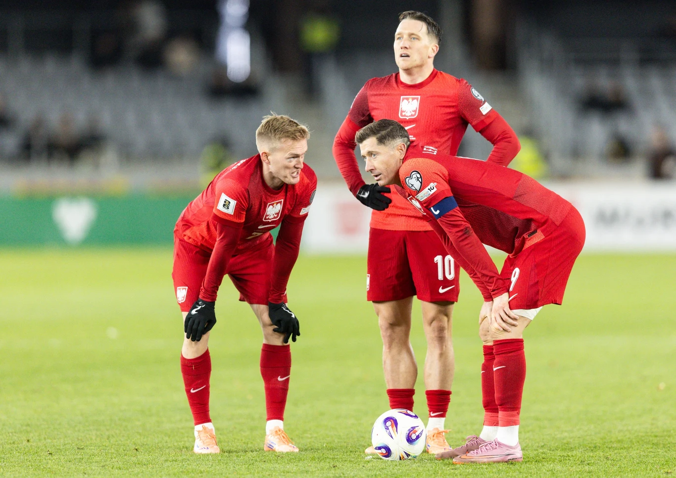 Karol Świderski, Piotr Zieliński i Robert Lewandowski w barwach reprezentacji Polski Trzech piłkarzy reprezentacji Polski w czerwonych strojach futbolowych przygotowuje się do wykonania rzutu wolnego na murawie stadionu, na pierwszym planie piłka, w tle trybuny.