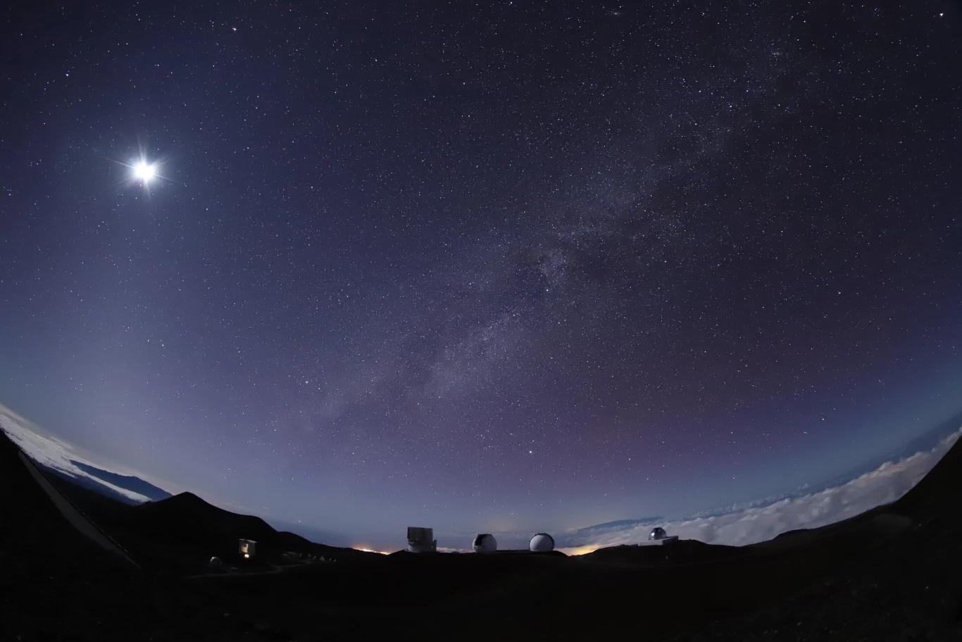 Odkrywca pierwszych planet spoza Układu Słonecznego uhonorowany planetoidą. Nocne niebo pokryte milionami gwiazd oraz jaśniejszy obiekt po lewej stronie kadru, najprawdopodobniej Księżyc, w dolnej części widoczne sylwetki kopuł obserwatoriów astronomicznych na tle gór oraz delikatnie podświetlonej linii horyzontu.