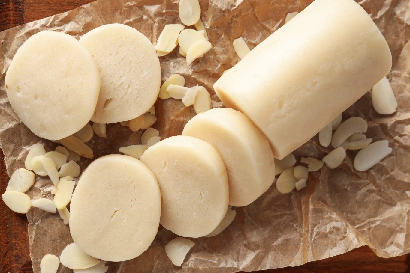 Pokrojony wałek marcepanu leży na brązowym papierze, w otoczeniu cienko pokrojonych płatków migdałowych.