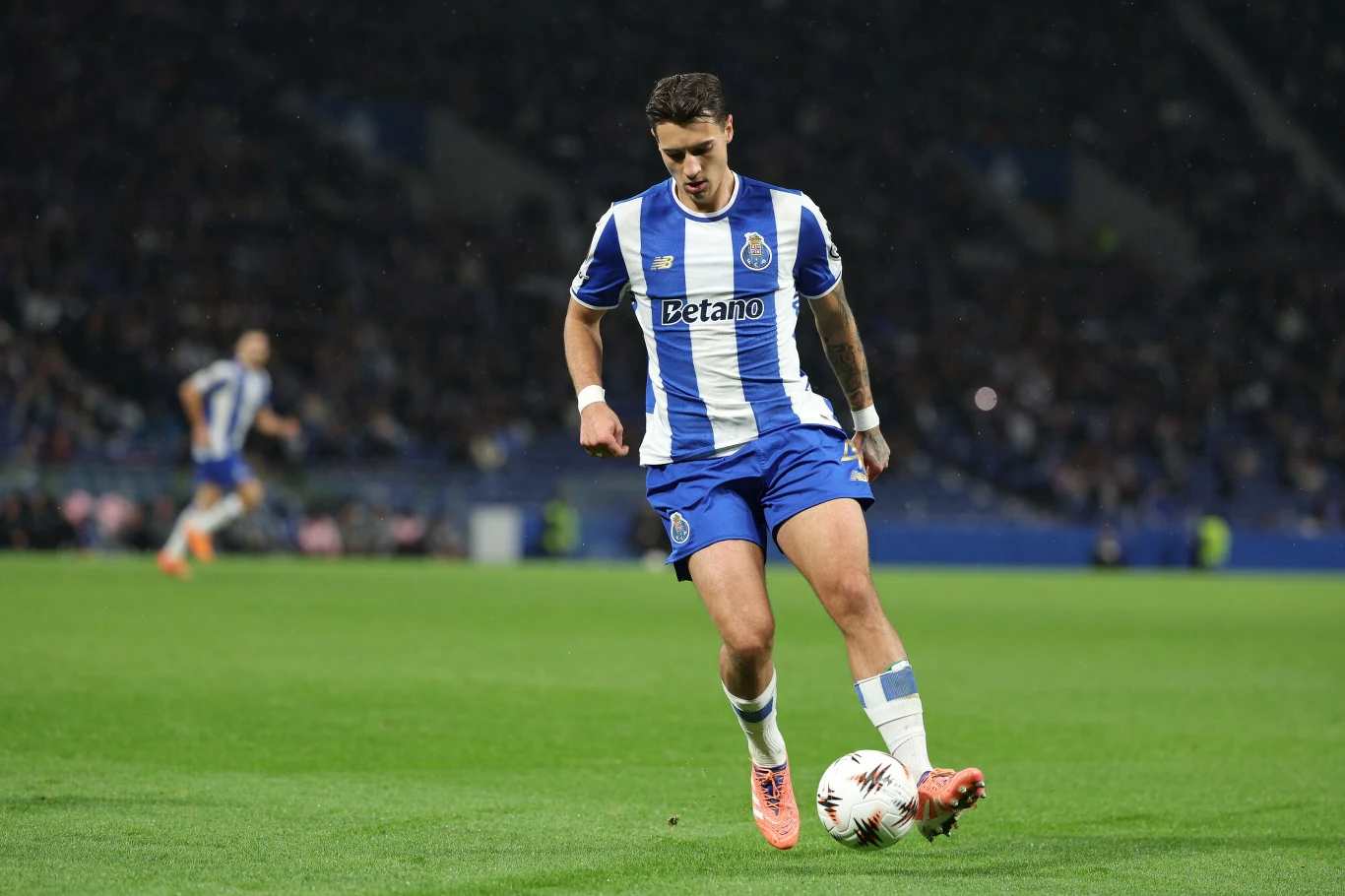 Jakub Kiwior Piłkarz w stroju biało-niebieskim prowadzi piłkę po zielonej murawie stadionu podczas meczu piłki nożnej, na trybunach widoczni widzowie.