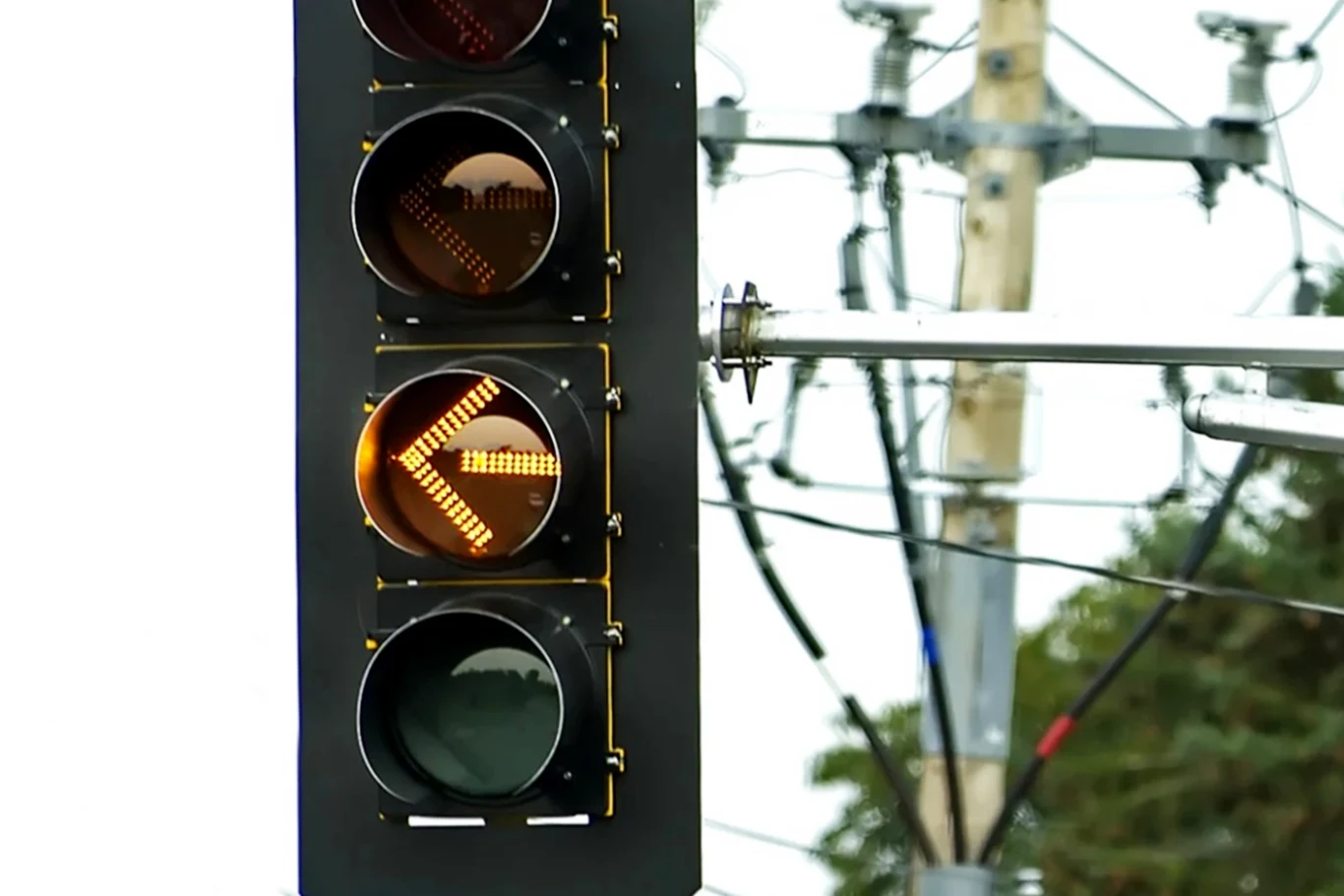 Żółta strzałka do skrętu w lewo. Wkrótce nowy sygnalizator? Sygnalizator świetlny z pomarańczową strzałką w lewo, w tle widoczne przewody i słup elektryczny oraz fragmenty drzew.