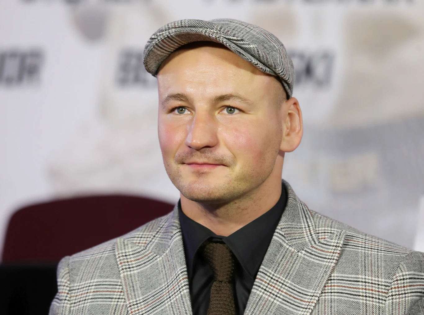 Mężczyzna w eleganckim kraciastym garniturze oraz płaskiej czapce, siedzący na tle rozmytego bannera z napisem, z wyraźnym uśmiechem na twarzy.