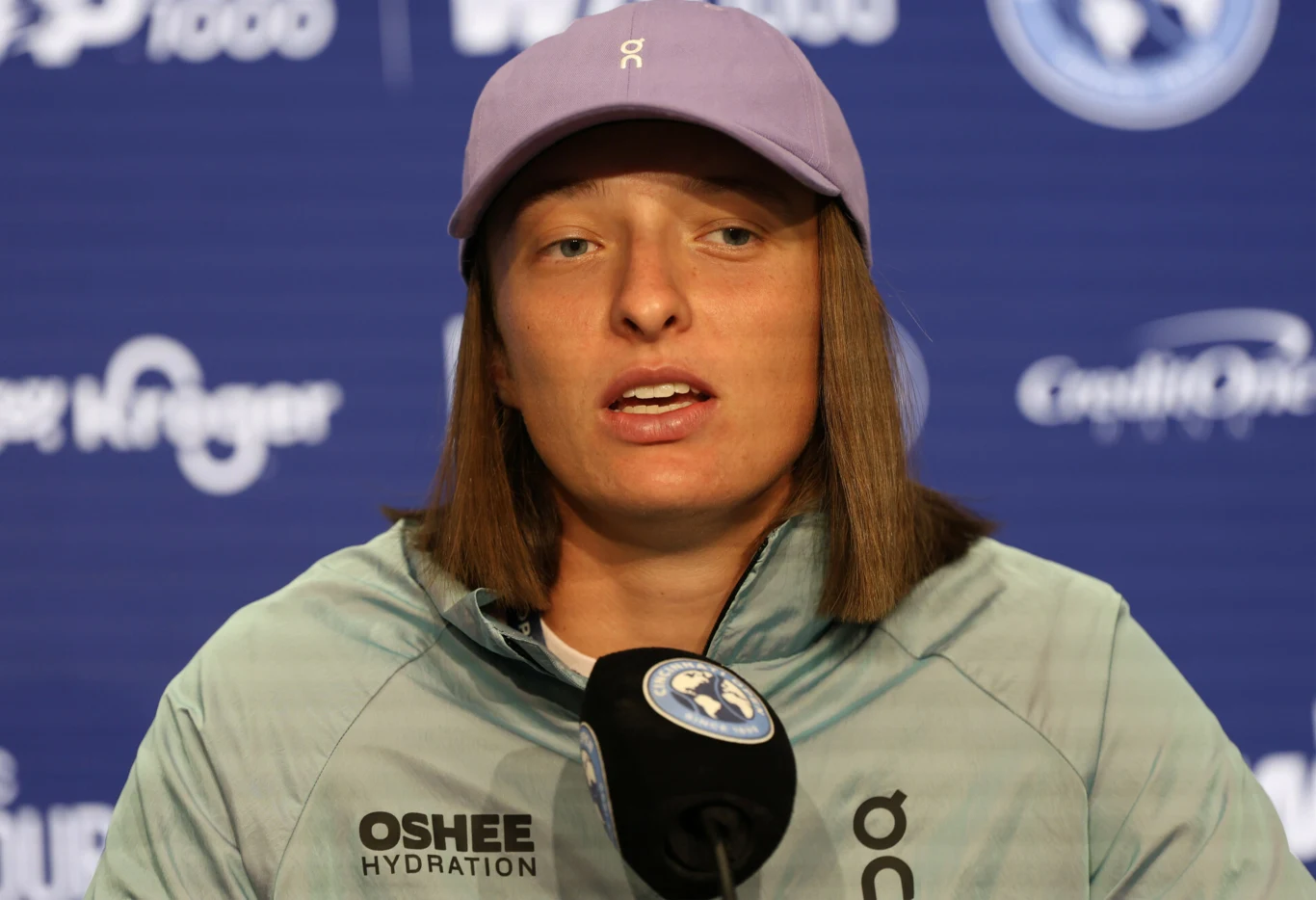 Kobieta w jasnej kurtce i fioletowej czapce z daszkiem siedzi przy mikrofonie podczas konferencji prasowej na tle tablicy z logotypami sponsorów i turnieju sportowego.