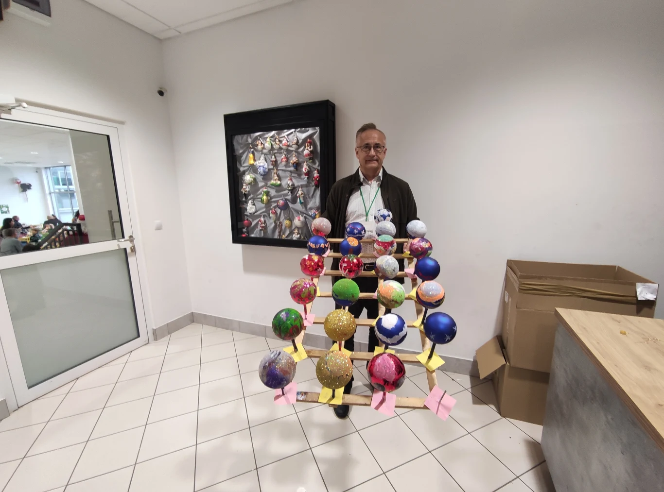 Marek Długosz z bombkami choinkowymi ozdobionymi przez dzieci podczas warsztatów zdobienia Mężczyzna w okularach stoi w jasnym pomieszczeniu, trzymając konstrukcję z kolorowych kul połączonych patyczkami, przypominającą model molekularny. W tle widoczny karton oraz wiszący na ścianie obraz. Na zewnątrz przez szybę widać ludzi.