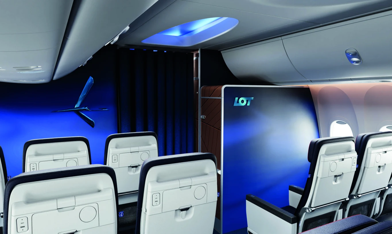 Wnętrze samolotu pasażerskiego LOT z rzędami wygodnych foteli, niebieskim oświetleniem oraz ozdobną przegrodą z logo przewoźnika.