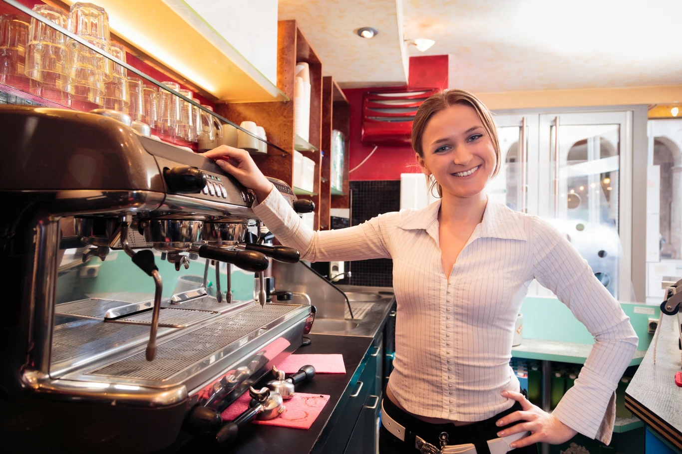 Młodzież może rozpocząć pracę zarobkową dopiero po ukończeniu 16. roku życia. Uśmiechnięta kobieta stojąca za ladą w kawiarni, opierająca się o dużą, profesjonalną maszynę do kawy. W tle półki z ustawionymi szklankami i jasne wnętrze lokalu.