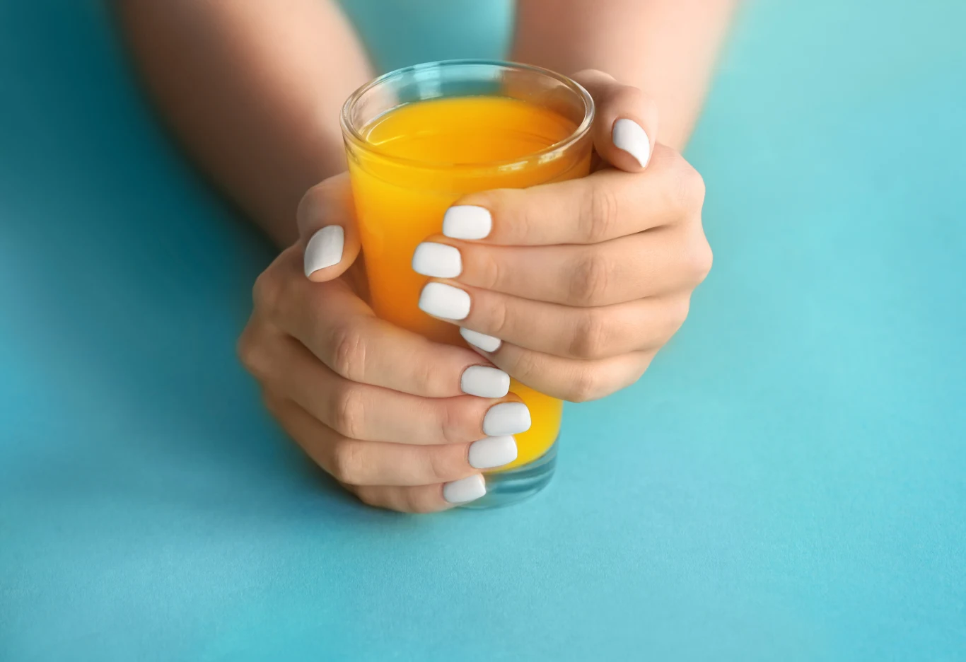 Dwie dłonie z białym manicure trzymają szklankę pełną pomarańczowego soku, tło jednolite, błękitne.