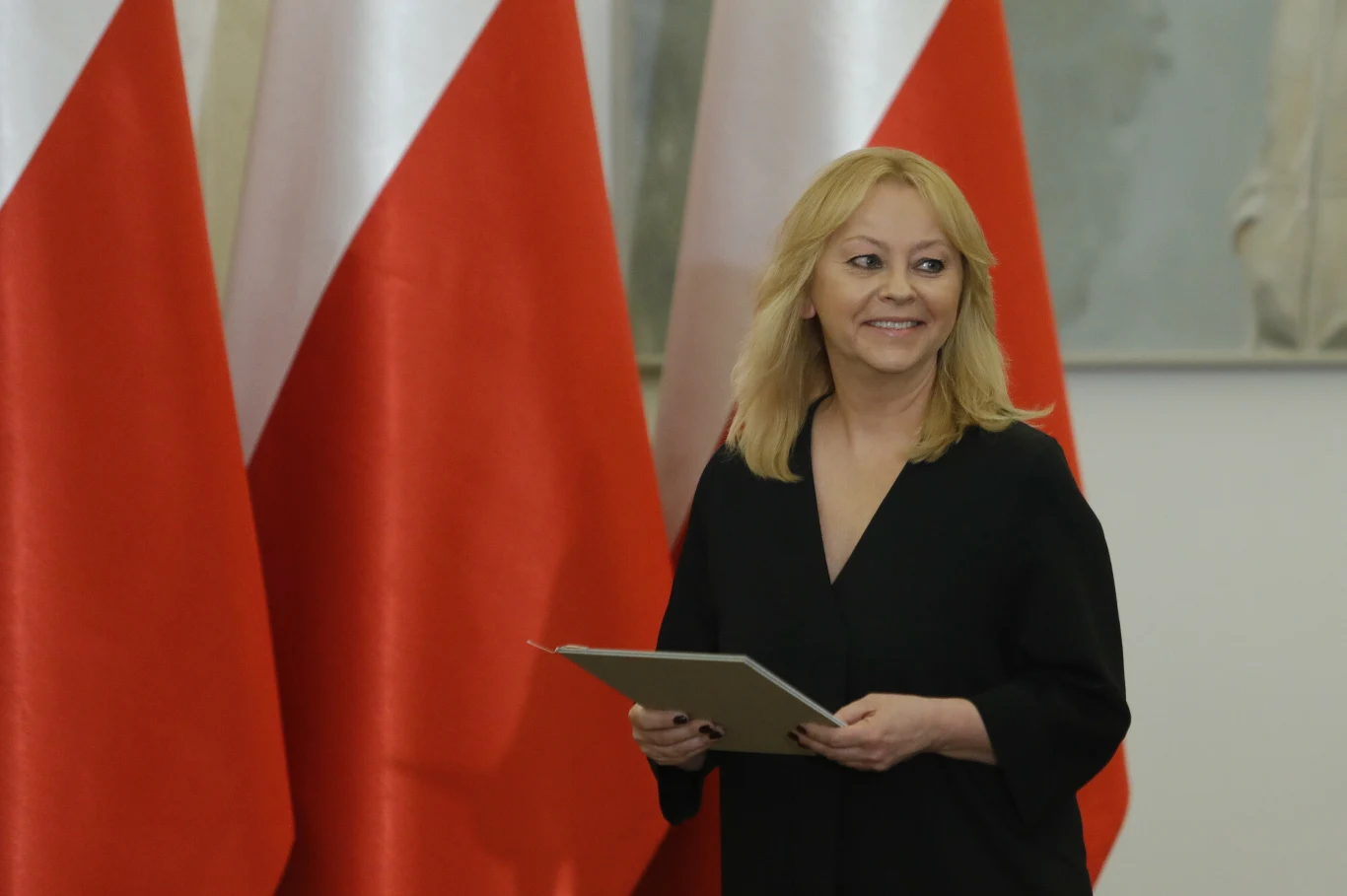 Kobieta z blond włosami ubrana na czarno trzyma dokument w dłoni, stojąc na tle trzech biało-czerwonych flag.