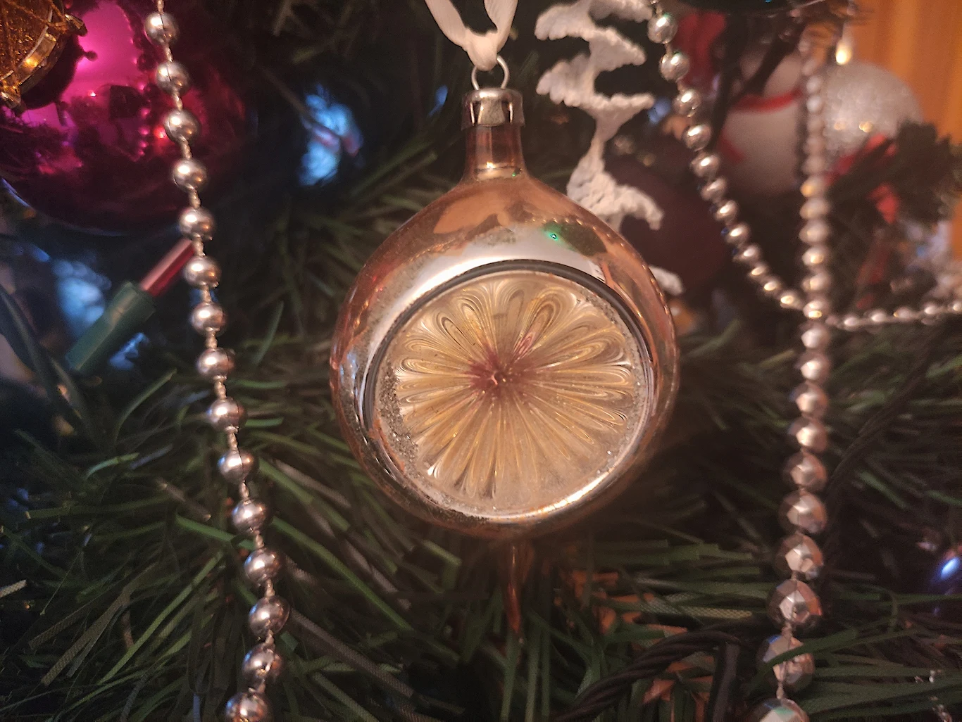 Bańka z reflektorem, popularna w PRL Szklana, okrągła bombka choinkowa z tłoczonym wzorem kwiatowym pośrodku, zawieszona na zielonej gałązce choinki. Bombkę otaczają srebrne koraliki jako dodatkowa ozdoba.