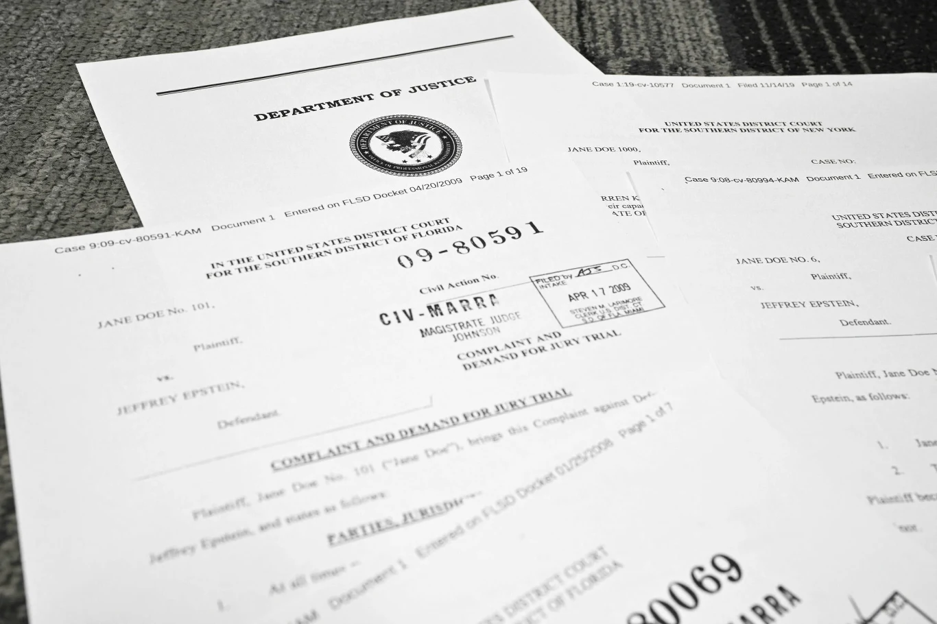 Rozłożone na szarej powierzchni oficjalne dokumenty sądowe Departamentu Sprawiedliwości Stanów Zjednoczonych związane ze sprawą przeciwko Jeffreyowi Epsteinowi, zawierające numery sprawy, daty oraz nazwiska stron.