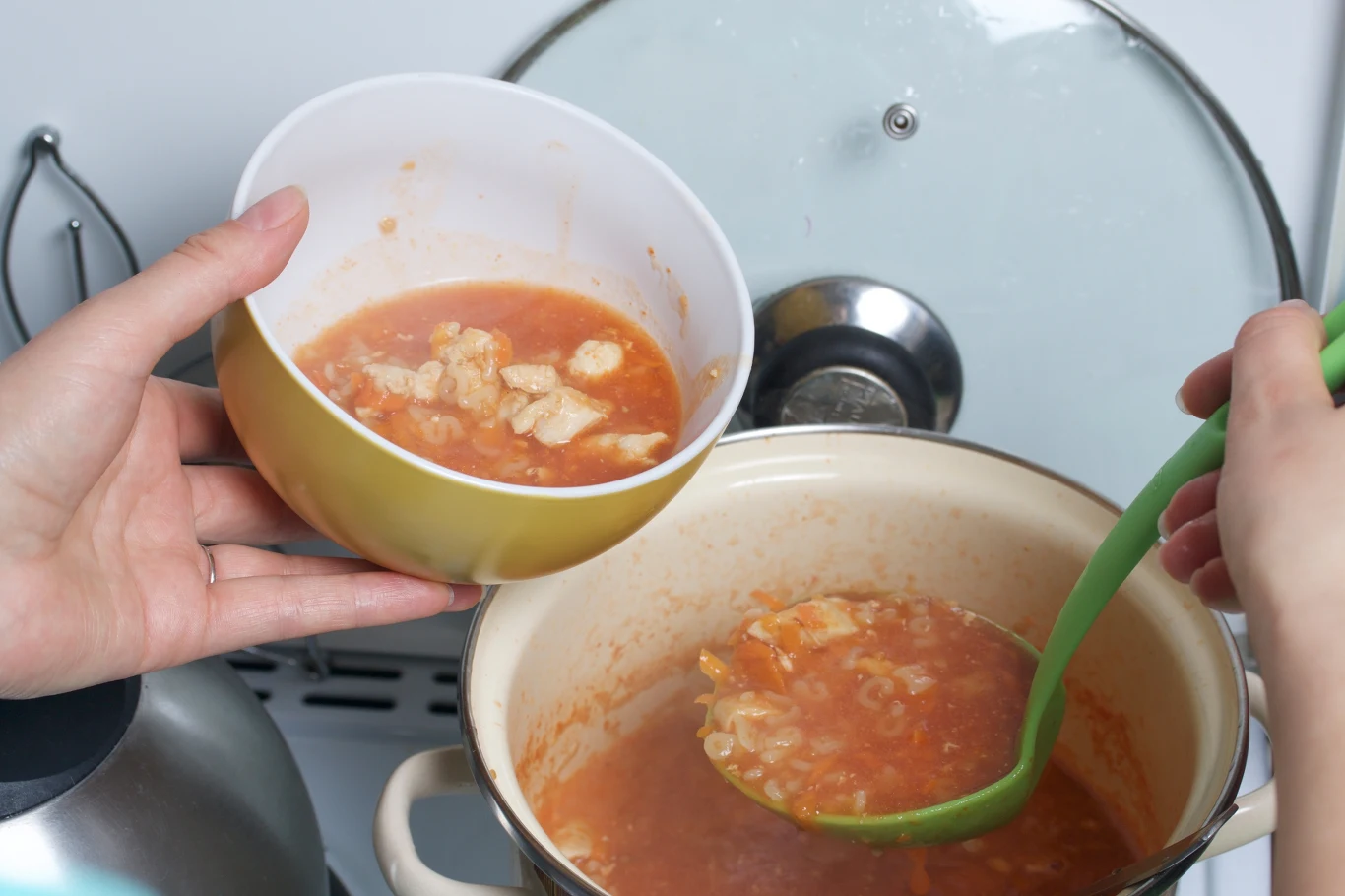 Jaką zupę warto zjeść po ciężkostrawnych potrawach? Gotowa do podania miska zupy pomidorowej z kawałkami mięsa, nabierana chochlą z garnka stojącego na kuchence.