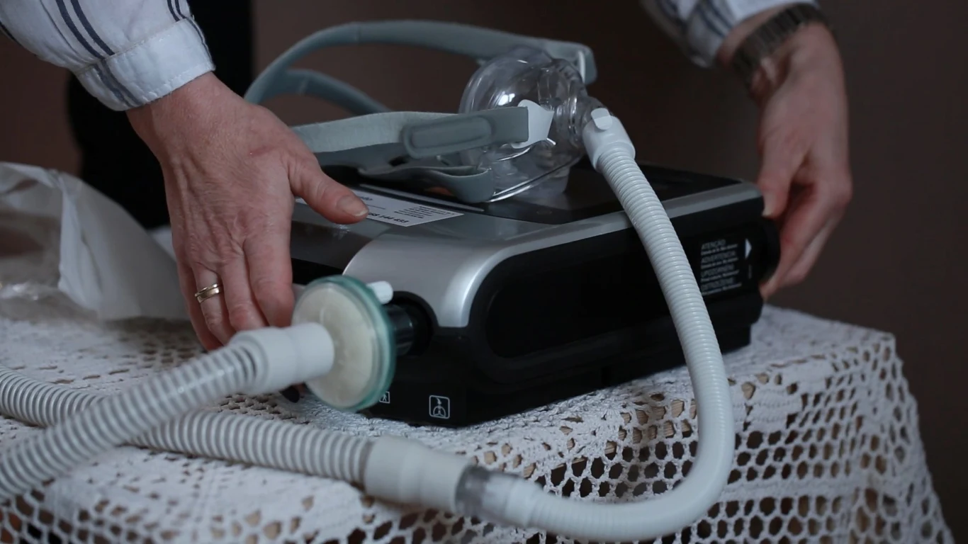 Ogólnopolski Związek Świadczeniodawców Wentylacji Mechanicznej Ręce osoby ustawiające na koronkowym obrusie urządzenie medyczne typu CPAP z dołączoną maską i rurkami, przeznaczone do terapii oddechowych.