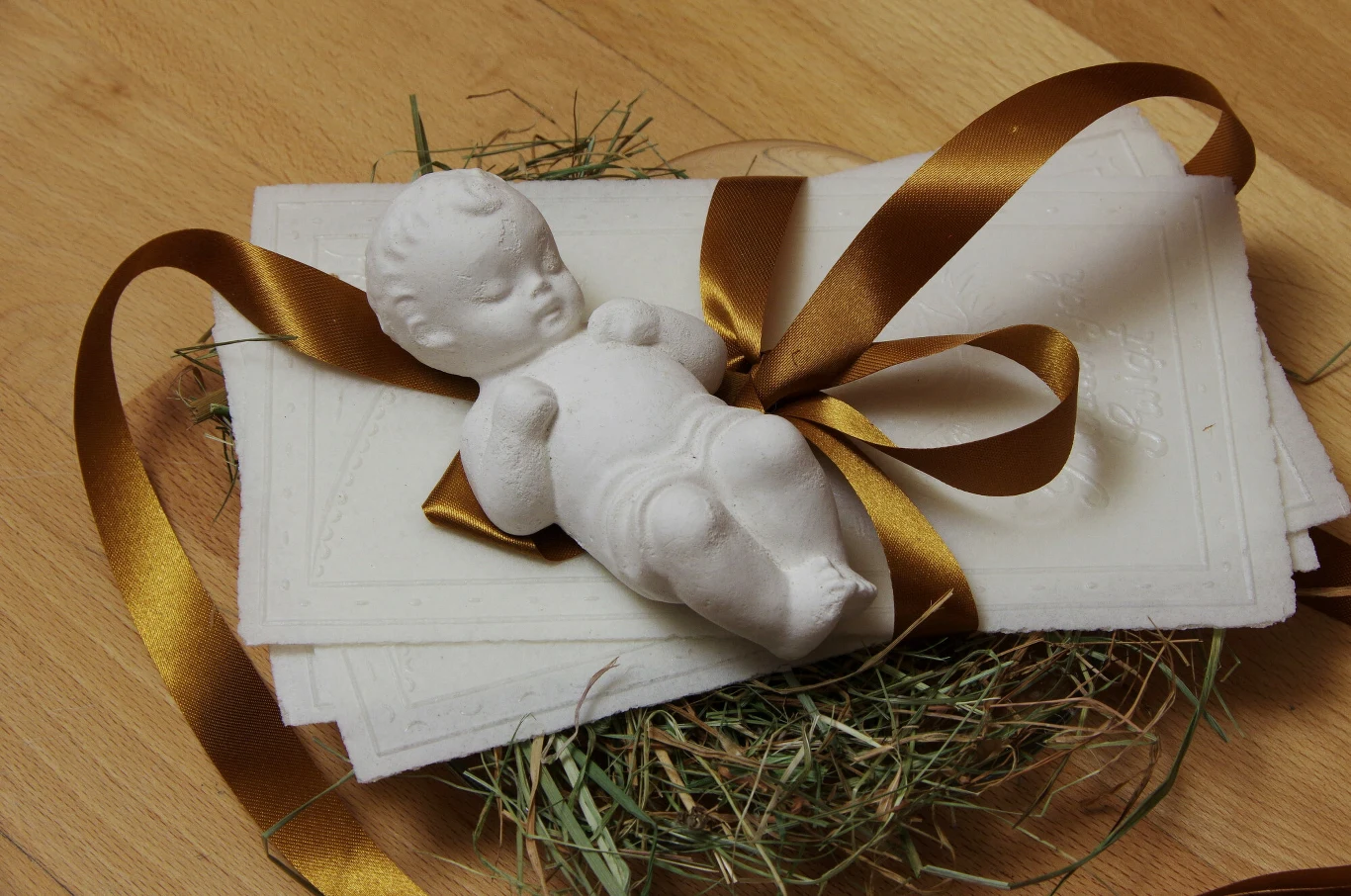 Opłatek na sianku, które tradycyjnie kładzie się pod obrus na wigilijnym stole Figurka niemowlęcia wykonana z białego materiału, przypominająca Dzieciątko Jezus, ułożona na stosie białych kart zawiązanych brązową wstążką, całość oparta na podłożu z suchej trawy, na drewnianym blacie.