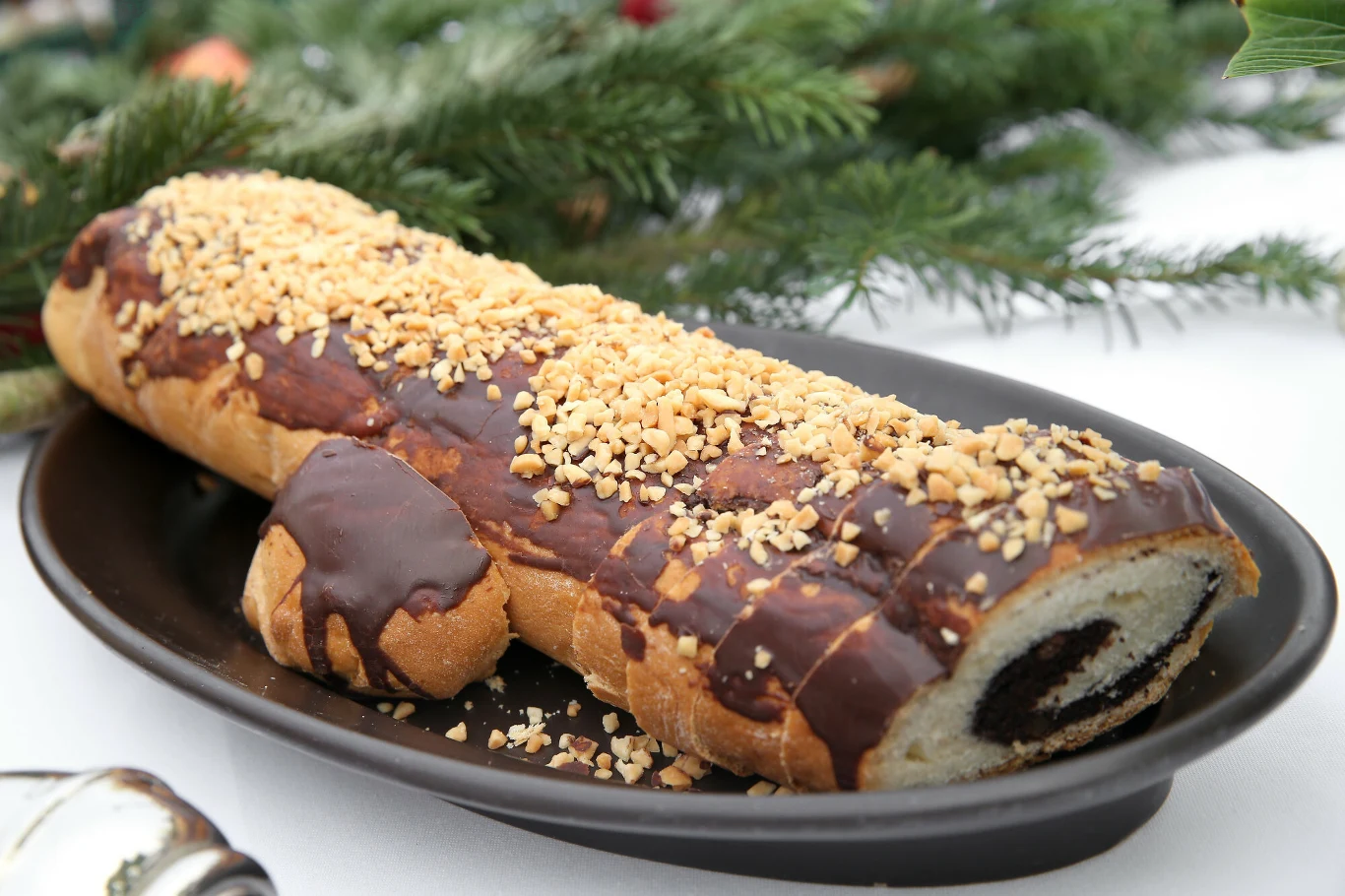 Tradycyjny świąteczny makowiec Rolada biszkoptowa pokryta polewą czekoladową i posypana drobno siekanymi orzechami, ułożona na ciemnym półmisku z zielonymi gałązkami iglastymi w tle.