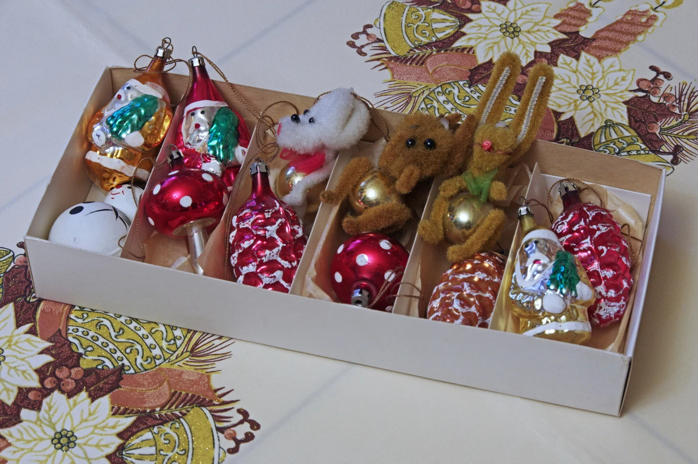Bombki z czasów PRL Kolorowe bombki choinkowe, wśród nich figurki Mikołaja, szyszki, zwierzątka z futerka i szklane ozdoby, ułożone w kartonowym pudełku na świątecznym obrusie.