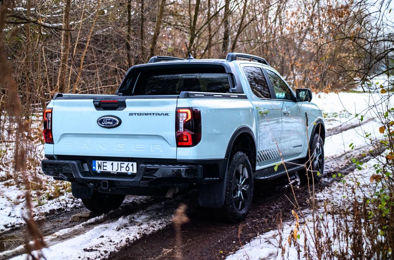 Jasnoniebieski pickup Ford Ranger Stormtrak stojący na leśnej, błotnistej drodze częściowo pokrytej śniegiem, otoczony drzewami bez liści.