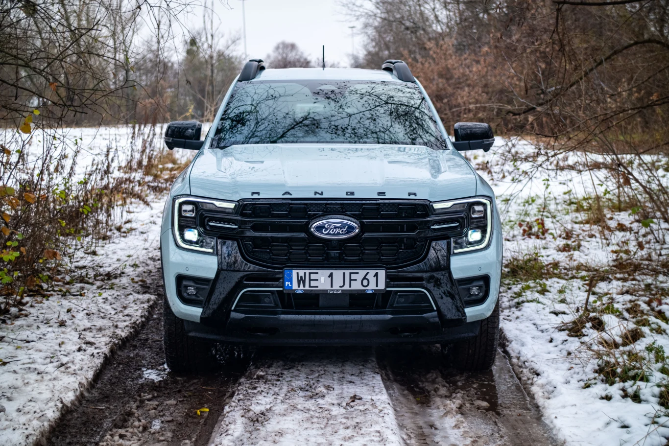 Biały Ford Ranger z czarnymi elementami, stojący na ośnieżonej, leśnej drodze. Teren dookoła to półnagie drzewa i krzewy, widoczne ślady opon w błocie. Samochód ustawiony centralnie, światła mijania włączone.